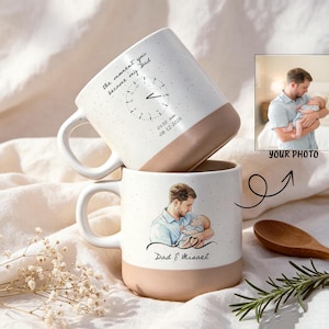 May include: Two ceramic mugs with a speckled white upper and solid tan base. One mug features a clock graphic with the text "the moment you became my Dad" and "03:20 am 08-12-2026". The other mug displays a custom illustration of a person holding a baby.