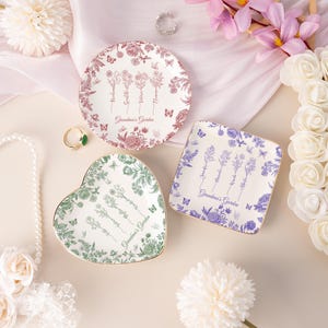 Pu&ograve; includere: Tre piatti decorativi in ceramica di diverse forme: rotondo, a cuore e quadrato. Ogni piatto presenta disegni floreali e il testo "Grandma's Garden". Il piatto rotondo ha un motivo floreale rosso, il piatto a forma di cuore ha un motivo floreale verde e il piatto quadrato ha un motivo floreale viola.