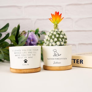 May include: Two white ceramic planters with bamboo saucers. One planter has the text "I LOVED YOU FOR YOUR WHOLE LIFE AND I'LL MISS YOU FOR THE REST OF MINE" and the name "COLIN". The other has a dog silhouette and the text "NEVER FORGOTTEN" and the name "Julian". A cactus with an orange flower sits in one.
