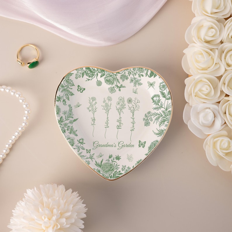 Pu&ograve; includere: Portagioie a forma di cuore con bordo dorato e design floreale verde. Il piatto presenta le parole "Grandma's Garden" e i nomi Faith, Victoria, Phyllis e Winifred. Farfalle e fiori decorano il piatto.
