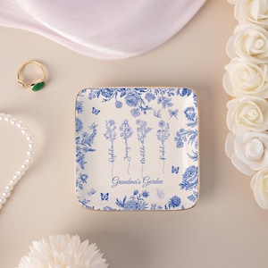 Pu&ograve; includere: Un vassoio quadrato in ceramica bianco sporco con un motivo floreale blu e bordi dorati. Il vassoio presenta le parole "Grandma's Garden" e i nomi Ophelia, Jenny, Matilda e Rachel. Le farfalle sono incluse nel design.