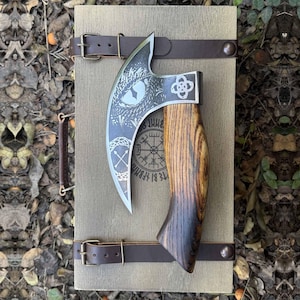 May include: A Viking-style axe with a curved blade featuring etched dragon and knotwork designs. The wooden handle has a dark stain, and the axe is secured to a wooden board with leather straps. The board has a leather handle and is tan.