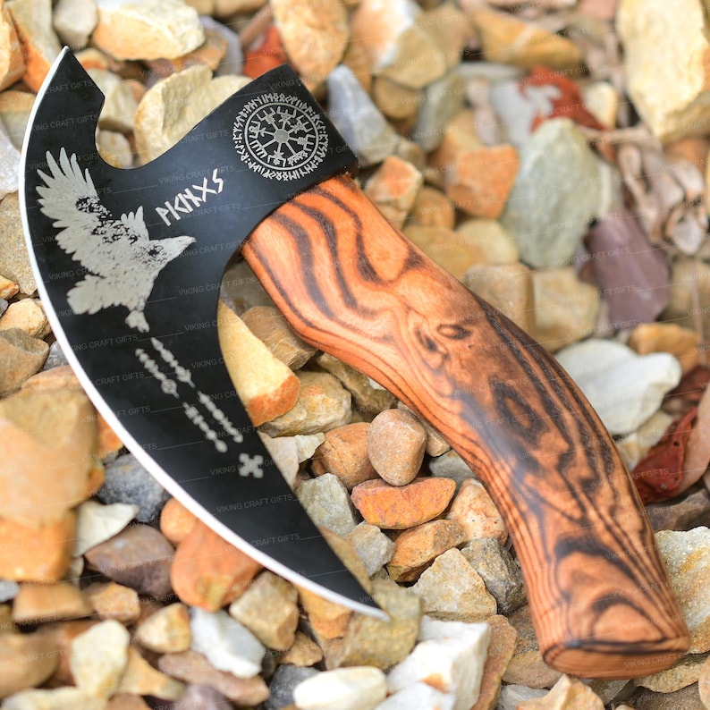 May include: A black and silver axe with a wooden handle. The axe has a Viking symbol and a raven design on the blade. The handle is made of dark wood with a wood grain pattern.