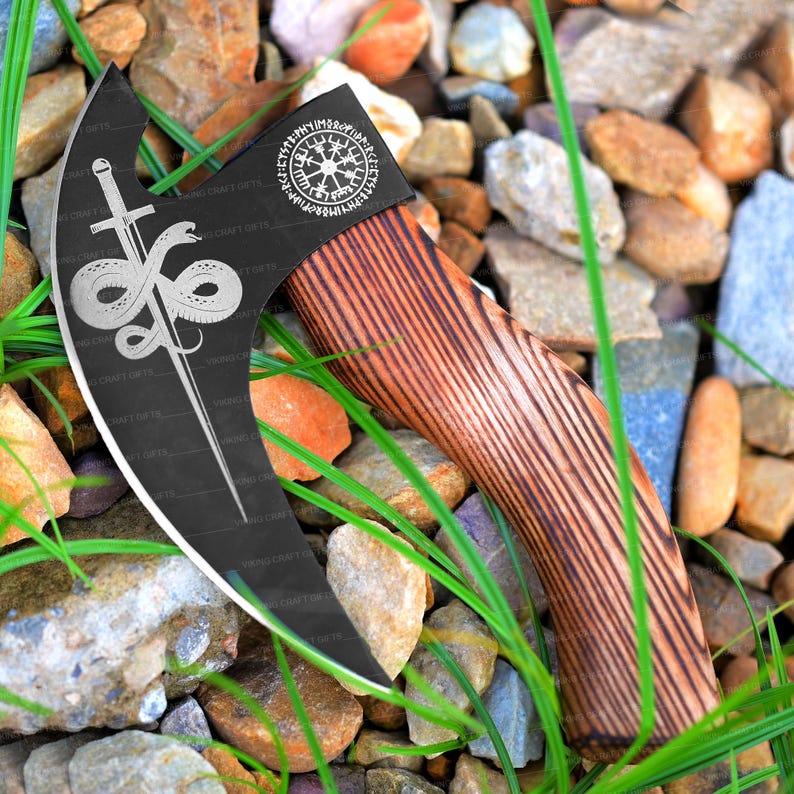 May include: A black axe with a wooden handle. The axe head has a silver design of a snake and a sword, and a circular compass with runes around it. The axe is resting on a bed of rocks and grass.