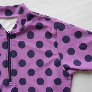 Peut inclure: Un chemisier à manches courtes de couleur violette avec une fermeture éclair blanche et un motif de pois bleu marine.