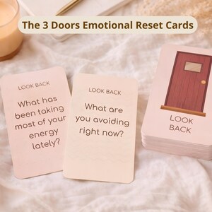 Puede incluir: Un juego de cartas con una ilustración de puerta de madera y las palabras "LOOK BACK" y "The 3 Doors Emotional Reset Cards". Dos cartas individuales muestran preguntas: "¿Qué te ha estado quitando la mayor parte de tu energía últimamente?" y "¿Qué estás evitando ahora mismo?"