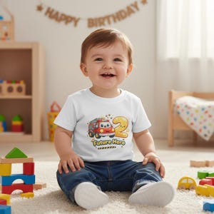 Op de afbeelding: Een lachende peuter draagt een wit t-shirt met een cartoon brandweerwagen, het nummer 2 en de woorden "Future Hero". Het kind zit op een crèmekleurig tapijt en draagt een blauwe spijkerbroek. Een banner met "Happy Birthday" is op de achtergrond.