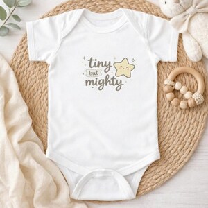 Può includere: Body da neonato bianco con la scritta "tiny but mighty" e una stella sorridente. Il body è appoggiato su un tappetino intrecciato, con un coniglietto di peluche e un anello per la dentizione in legno sullo sfondo.