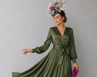 May include: A long-sleeved olive green satin wrap dress with a V-neckline and a tie waist. The dress features a full, flowing skirt. The ensemble is paired with a large floral headpiece in shades of purple and green, and bright pink heels and clutch.
