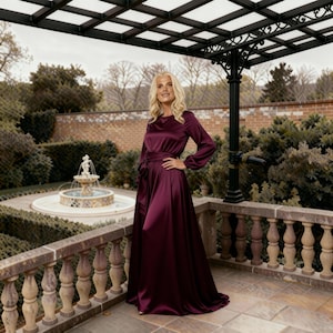 May include: A full-length, burgundy satin dress with long sleeves and a belted waist. The dress is worn outdoors on a stone patio with a decorative railing, with a fountain and manicured garden in the background.