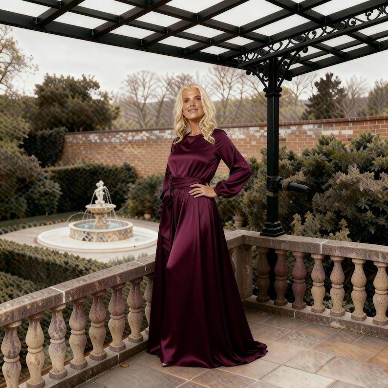 May include: A full-length, burgundy satin dress with long sleeves and a belted waist. The dress is worn outdoors on a stone patio with a decorative railing, with a fountain and manicured garden in the background.