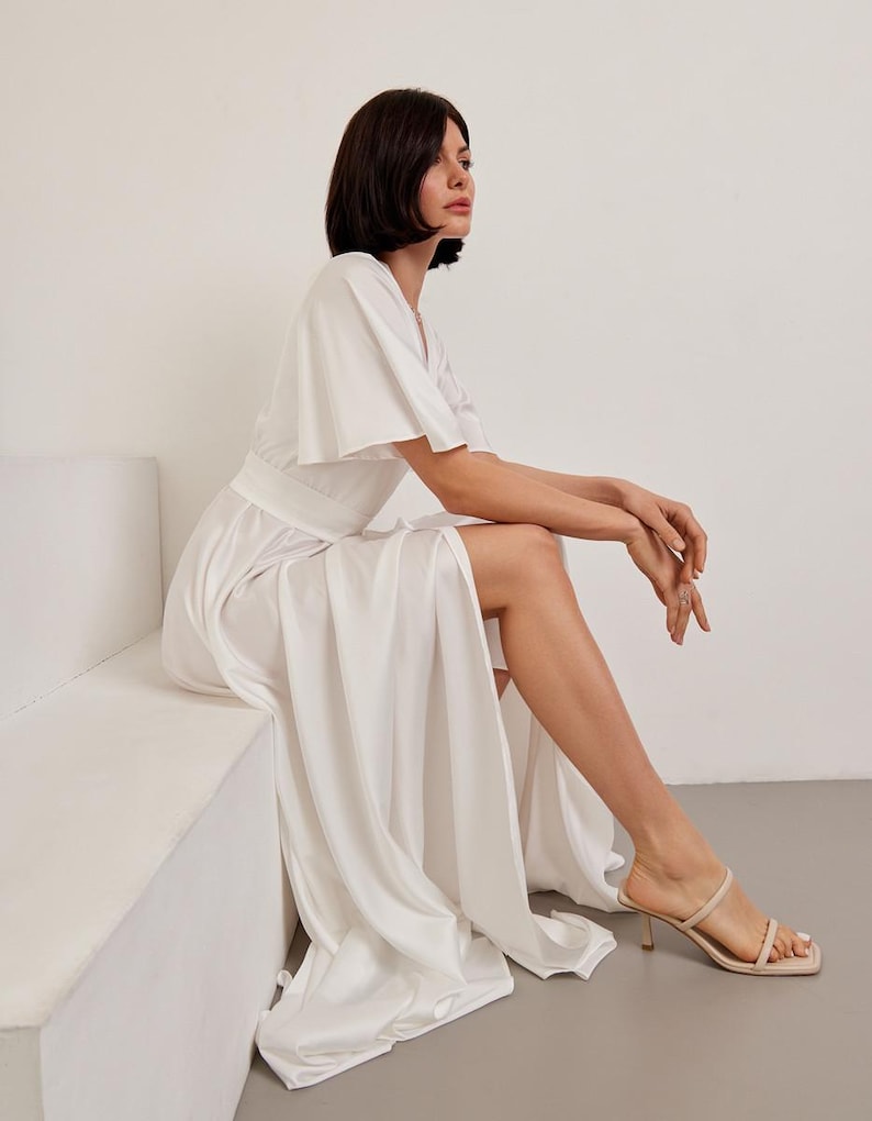 May include: A white, flowing dress with short, flared sleeves and a high slit. The dress is paired with beige heeled sandals. The model is seated on a white surface, with a neutral background.