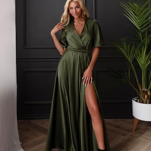 May include: An olive green satin wrap dress with short sleeves and a high slit. The dress is paired with black heeled sandals. A woman is wearing the dress. A potted plant is in the background.