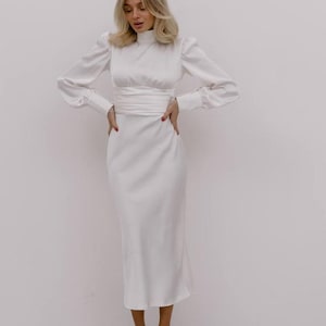 May include: A white midi-length dress featuring a high mock neckline, long sleeves with buttoned cuffs, and a wide, wrapped waist detail. The garment is constructed from a smooth, satin-like fabric that falls to a straight, clean-lined skirt hem.