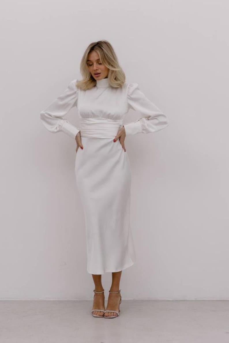 May include: A white midi-length dress featuring a high mock neckline, long sleeves with buttoned cuffs, and a wide, wrapped waist detail. The garment is constructed from a smooth, satin-like fabric that falls to a straight, clean-lined skirt hem.