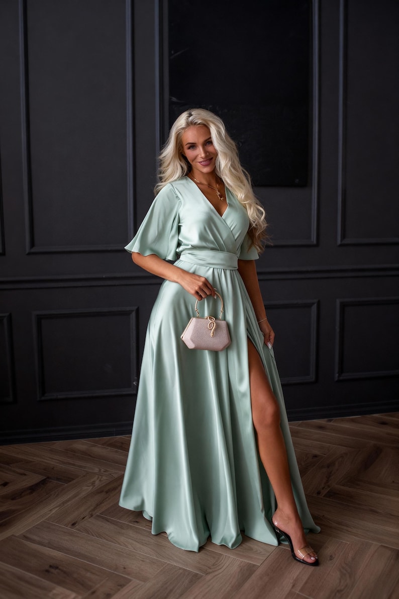 May include: A long, flowing, mint green satin dress with a V-neckline and short sleeves. The dress features a high slit and is paired with a small, light pink purse. The background is a dark wall and wooden floor.
