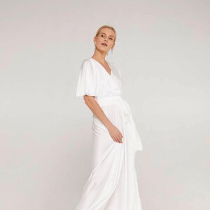 May include: A white wrap dress with short sleeves and a flowing skirt. The dress features a V-neckline and a waist tie. The model is wearing the dress in a studio setting with a neutral background. The dress is made of a soft, silky fabric.