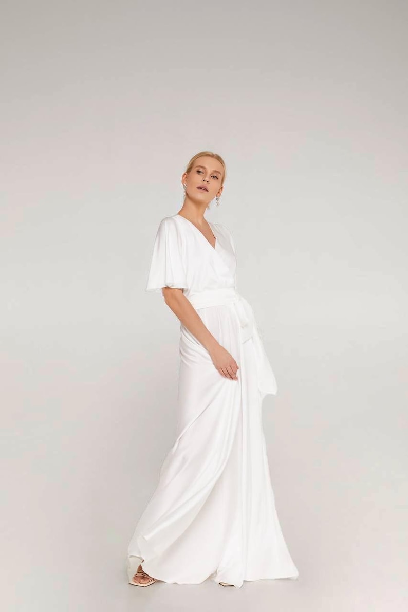 May include: A white wrap dress with short sleeves and a flowing skirt. The dress features a V-neckline and a waist tie. The model is wearing the dress in a studio setting with a neutral background. The dress is made of a soft, silky fabric.