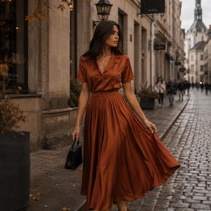 May include: A rust-colored satin two-piece set consisting of a short-sleeved button-down shirt with a collar and a matching pleated midi skirt. The outfit is paired with brown pointed-toe heels and a small black structured handbag.
