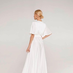 May include: A white, flowing dress with short sleeves and a cinched waist. The dress has a V-neckline and a long, flared skirt. The model is seen from the back, with blonde hair styled in a bun. The dress appears to be made of a soft, silky fabric.