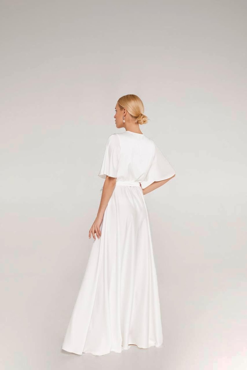 May include: A white, flowing dress with short sleeves and a cinched waist. The dress has a V-neckline and a long, flared skirt. The model is seen from the back, with blonde hair styled in a bun. The dress appears to be made of a soft, silky fabric.