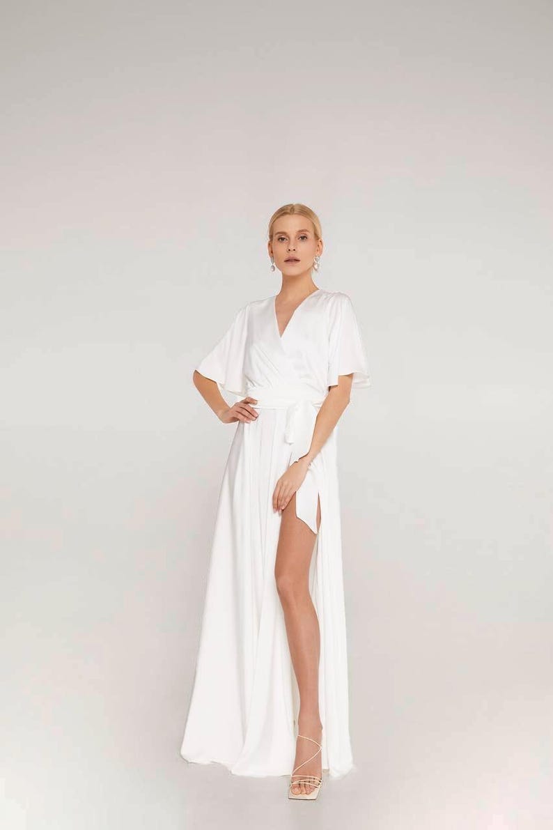May include: A white wrap dress with short sleeves and a high slit. The dress has a V-neckline and a matching waist tie. The model is wearing strappy sandals. The dress is made of a flowing fabric.