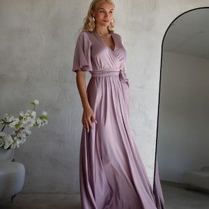May include: A flowing, mauve-colored dress with short, flutter sleeves and a wrap-style bodice. The dress features a waist tie and a floor-length skirt. The fabric has a subtle sheen, suggesting a satin or silk material. The dress is suitable for formal events.