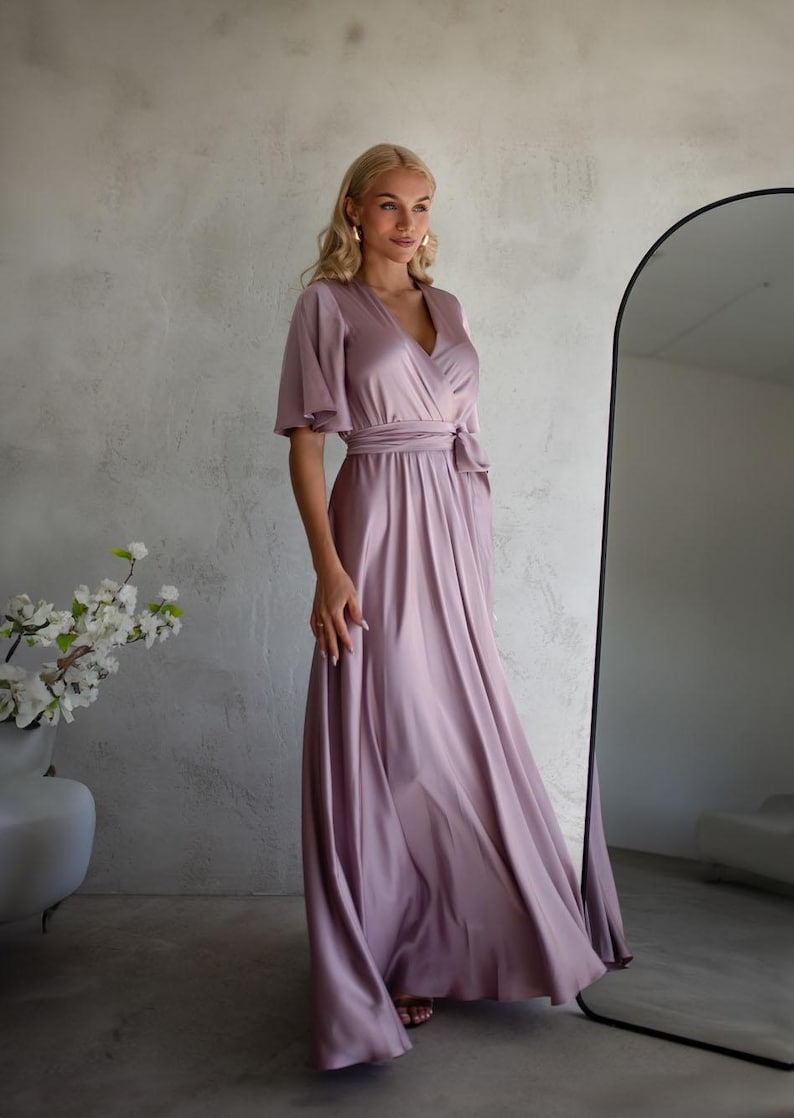 May include: A flowing, mauve-colored dress with short, flutter sleeves and a wrap-style bodice. The dress features a waist tie and a floor-length skirt. The fabric has a subtle sheen, suggesting a satin or silk material. The dress is suitable for formal events.