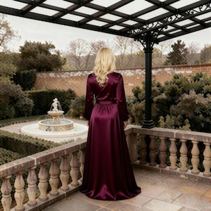 May include: A long, flowing, burgundy satin robe with long sleeves and a matching belt. The robe is worn outdoors on a stone patio overlooking a garden with a fountain. The robe's fabric has a subtle sheen.