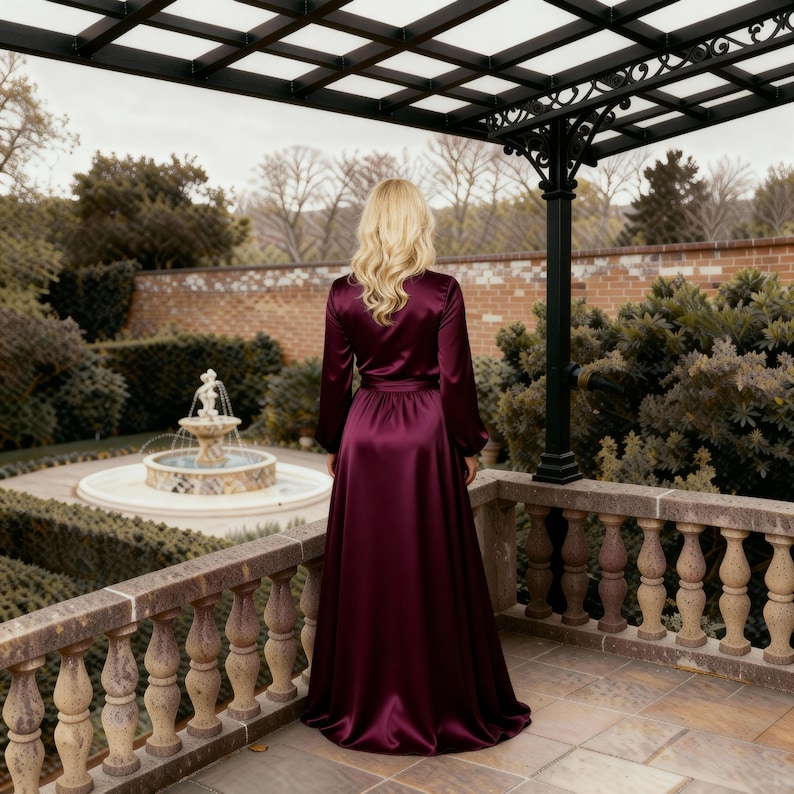 May include: A long, flowing, burgundy satin robe with long sleeves and a matching belt. The robe is worn outdoors on a stone patio overlooking a garden with a fountain. The robe's fabric has a subtle sheen.