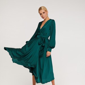 Puede incluir: Un vestido largo cruzado verde esmeralda con mangas largas y cintur&oacute;n anudado. El vestido tiene un escote en V y una falda fluida. La modelo lleva sandalias blancas. El vestido es de un material brillante y sedoso.