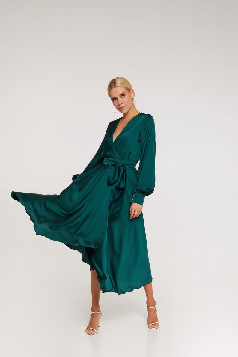 Puede incluir: Un vestido largo cruzado verde esmeralda con mangas largas y cintur&oacute;n anudado. El vestido tiene un escote en V y una falda fluida. La modelo lleva sandalias blancas. El vestido es de un material brillante y sedoso.
