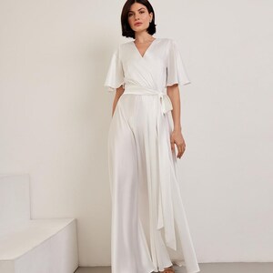 May include: A white jumpsuit with a wrap-style bodice and wide-leg pants. The jumpsuit features short, flutter sleeves and a matching fabric belt that ties at the waist. The model is wearing gold sandals.