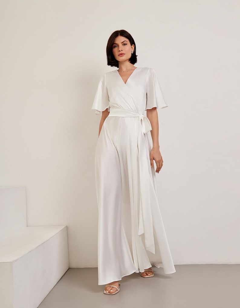 May include: A white jumpsuit with a wrap-style bodice and wide-leg pants. The jumpsuit features short, flutter sleeves and a matching fabric belt that ties at the waist. The model is wearing gold sandals.