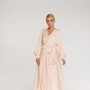 May include: A light peach-colored wrap dress with long sleeves and a midi-length skirt. The dress features a V-neckline, a tie-waist, and a flowing skirt. The sleeves have a slight puff at the shoulder. The model is wearing white high-heeled sandals.