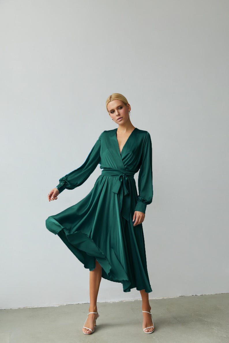 Puede incluir: Vestido cruzado verde esmeralda de manga larga, cuello en V y cintur&oacute;n a juego. El vestido tiene una falda fluida y est&aacute; hecho de un material brillante y sedoso. La modelo lleva sandalias blancas.