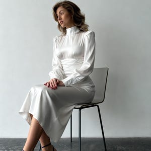 May include: A long-sleeved white satin midi dress featuring a high mock neckline, gathered waist detailing, and cuffed sleeves. The dress has a smooth, lustrous finish and a flowing skirt that falls below the knees, paired with dark purple pointed-toe heels.
