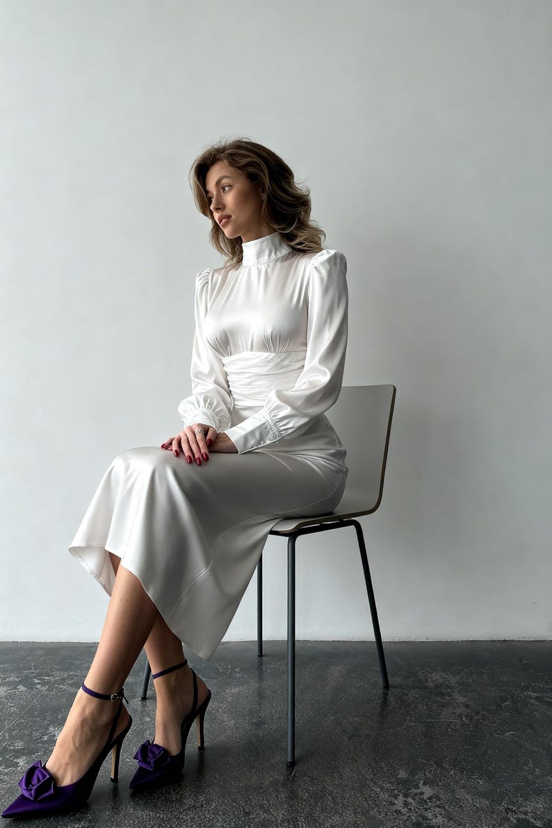 May include: A long-sleeved white satin midi dress featuring a high mock neckline, gathered waist detailing, and cuffed sleeves. The dress has a smooth, lustrous finish and a flowing skirt that falls below the knees, paired with dark purple pointed-toe heels.