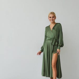 Puede incluir: Vestido cruzado verde oliva con mangas largas y una abertura alta. El vestido presenta un escote en V y un cintur&oacute;n de tela a juego atado a la cintura. La modelo lleva sandalias blancas.