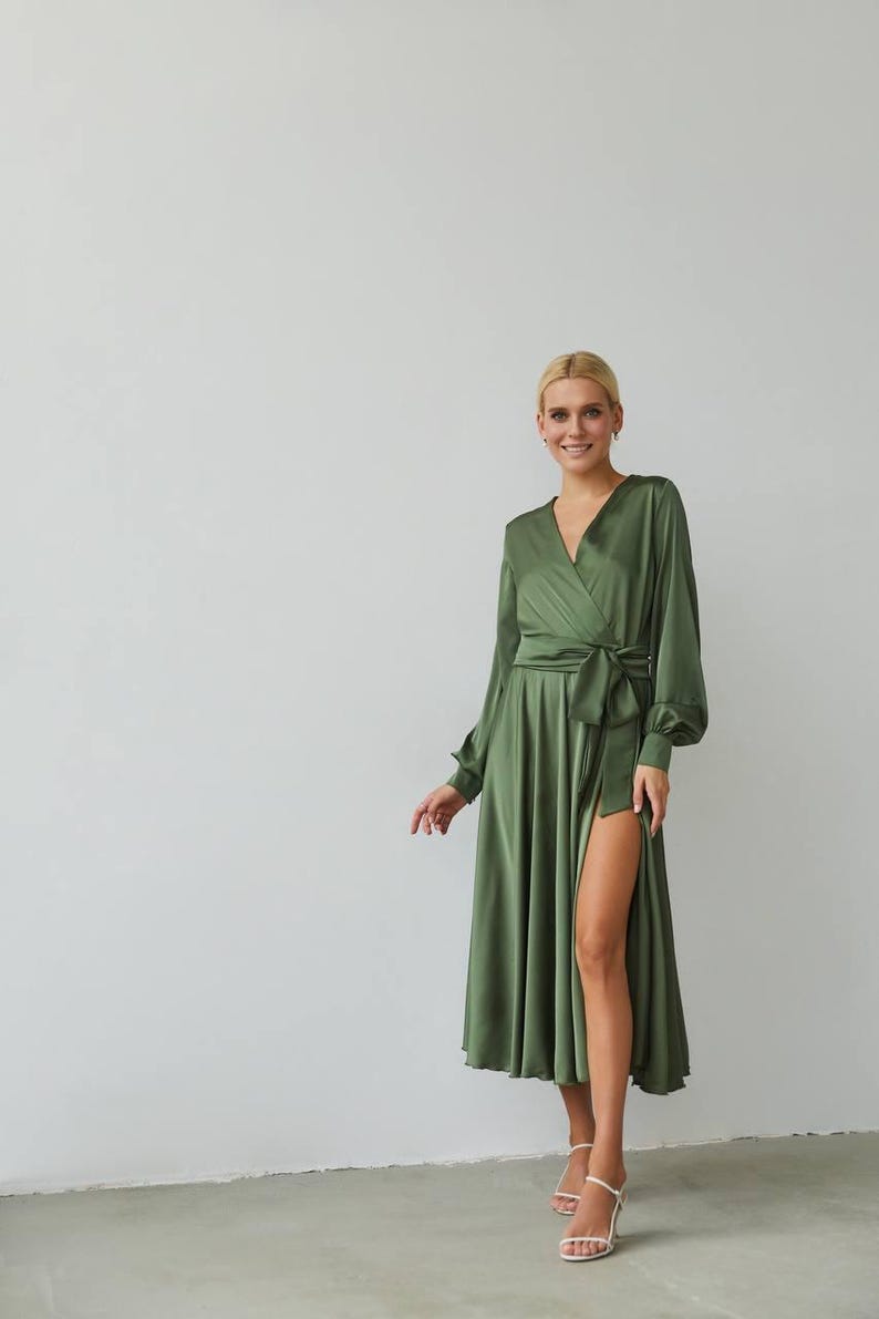 Puede incluir: Vestido cruzado verde oliva con mangas largas y una abertura alta. El vestido presenta un escote en V y un cintur&oacute;n de tela a juego atado a la cintura. La modelo lleva sandalias blancas.