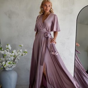 May include: A mauve-colored wrap dress with short, flutter sleeves and a high slit. The dress features a waist tie and is made of a flowing, silky material. A large vase with white flowers is in the background.