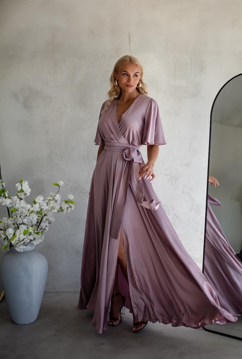 May include: A mauve-colored wrap dress with short, flutter sleeves and a high slit. The dress features a waist tie and is made of a flowing, silky material. A large vase with white flowers is in the background.