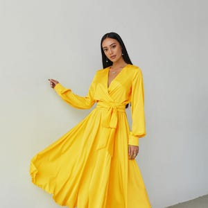 Puede incluir: Un vestido cruzado amarillo brillante con mangas largas y cintur&oacute;n anudado. El vestido tiene un escote en V y una falda fluida. La modelo lleva sandalias blancas con tiras. El vestido est&aacute; hecho de un material brillante y sedoso.