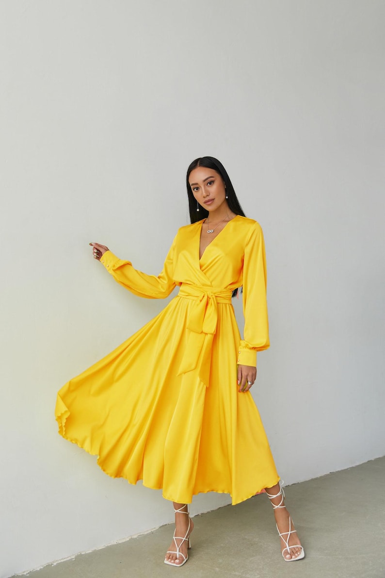 Puede incluir: Un vestido cruzado amarillo brillante con mangas largas y cintur&oacute;n anudado. El vestido tiene un escote en V y una falda fluida. La modelo lleva sandalias blancas con tiras. El vestido est&aacute; hecho de un material brillante y sedoso.
