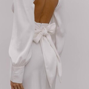 May include: A long-sleeved white dress featuring a high neckline and an open back design. The garment includes a large, prominent fabric bow tied at the waist, long sleeves with buttoned cuffs, and a smooth, solid white finish throughout the piece.