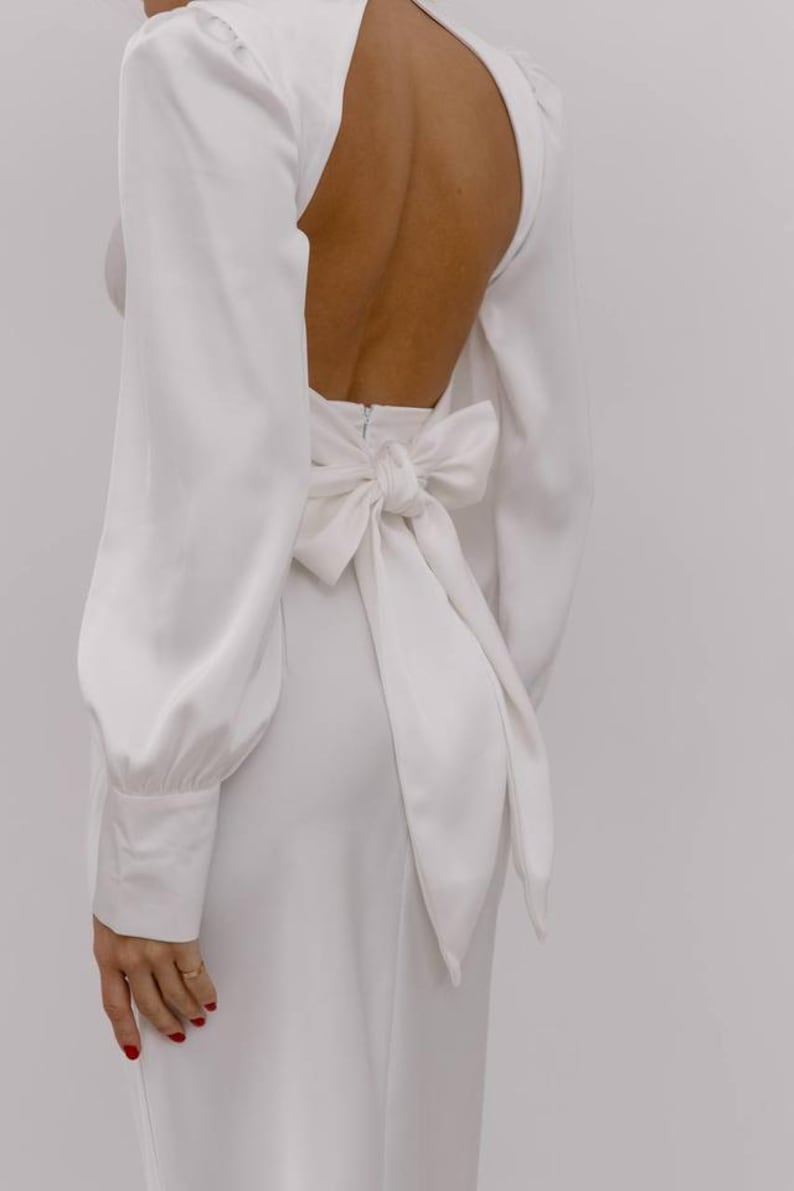 May include: A long-sleeved white dress featuring a high neckline and an open back design. The garment includes a large, prominent fabric bow tied at the waist, long sleeves with buttoned cuffs, and a smooth, solid white finish throughout the piece.