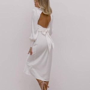 May include: A long-sleeved white midi dress featuring a high neckline, a large open back, and a prominent fabric bow tied at the waist. The dress has long sleeves with fitted cuffs and a smooth, solid white finish that falls to a mid-calf length.