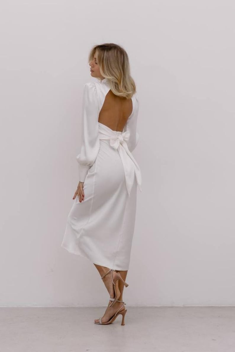 May include: A long-sleeved white midi dress featuring a high neckline, a large open back, and a prominent fabric bow tied at the waist. The dress has long sleeves with fitted cuffs and a smooth, solid white finish that falls to a mid-calf length.