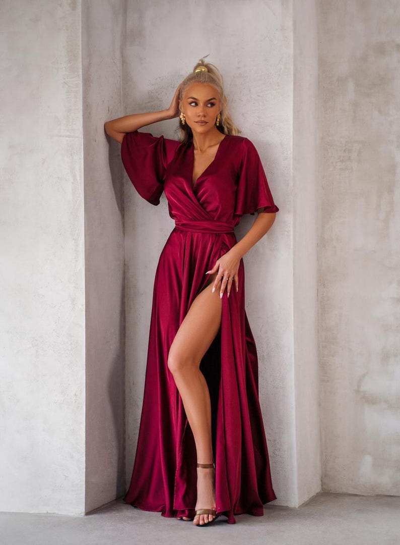 May include: A burgundy satin wrap dress with a high slit and short, flared sleeves. The dress features a V-neckline and a matching waist tie. The model is wearing brown sandals. The dress is floor-length.
