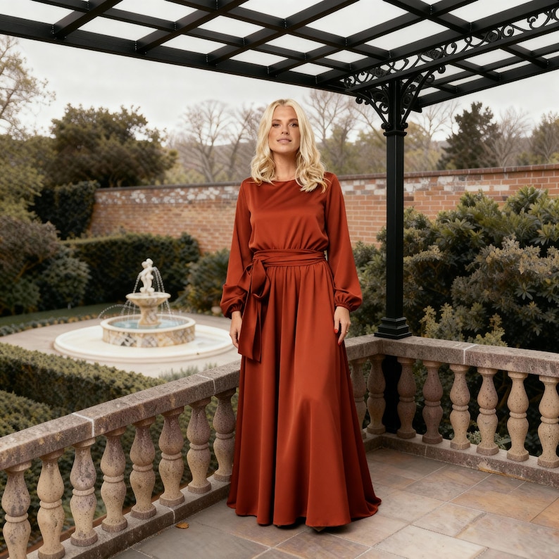 May include: A full-length, rust-coloured dress with long sleeves and a matching waist tie. The dress features a flowing skirt and is made of a shiny, satin-like material. The model is standing on a stone balcony with a fountain and garden in the background.